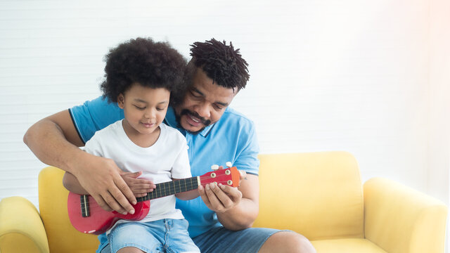 African dad teaching son how to play guitar on the sofa in the living room at home. Father's day, family bonding time concept.