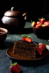 A piece of chocolate cake on a plate with strawberry slices and mint leaves on top. An earthenware plate, a mug, and a teapot of tea stand side by side on a dark table
