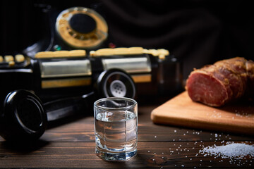 A vodka glass on an old table with an old black phone meat on the board with salt with shallow depth of field and blur.