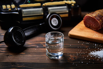 A vodka glass on an old table with an old black phone meat on the board with salt with shallow depth of field and blur.