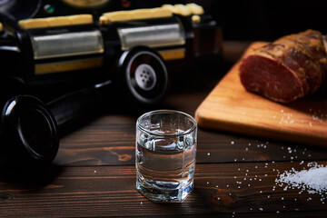 A vodka glass on an old table with an old black phone meat on the board with salt with shallow...