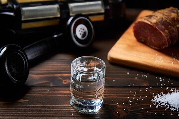 A vodka glass on an old table with an old black phone meat on the board with salt with shallow depth of field and blur.