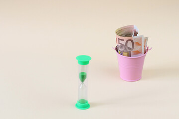 Light green, hourglass. Defocused money in a small pink bucket. Copying space is a concept of economy, crisis, business, savings, support.