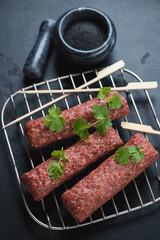 Metal rack with raw marbled beef lula kebabs, vertical shot over black stone surface, selective focus
