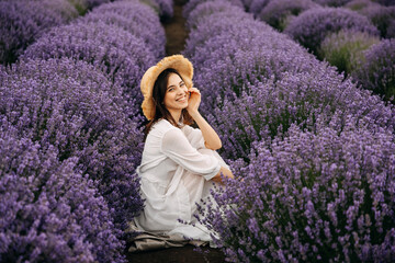 Young woman sitting between rows of lavender bushes, wearing a bohemian white dress and a straw hat, looking to camera.