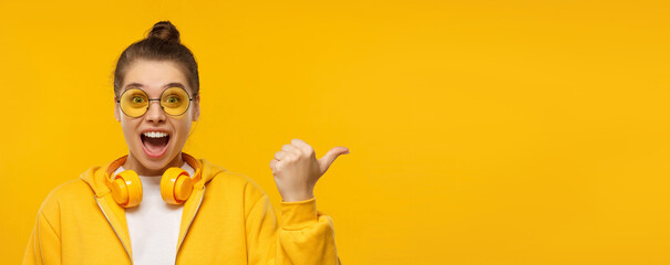 Horizontal banner of young excited girl wearing hoodie, glasses and wireless headphones around neck, pointing right to copy space, isolated on yellow background - obrazy, fototapety, plakaty