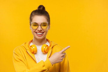 Young laughing girl wearing headphones around neck, pointing right, isolated on yellow background