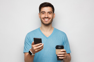Young man in and blue t-shirt holding coffee cup in one hand and smartphone in another, isolated on gray background