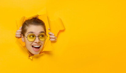 Horizontal banner of young girl in glasses tearing paper and peeking out hole, curious about...