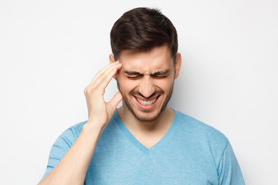 Headache concept. Young man touching temple with hand, trying to relieve pain, experiencing severe migraine, isolated on gray background