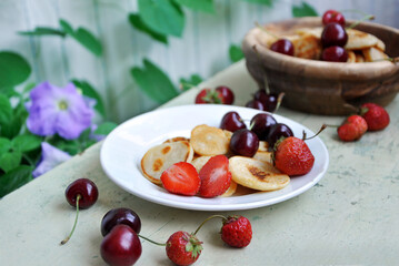 Homemade Mini pancakes and fresh strawberries, cherries on an old wooden table. Garden flowers in the background.