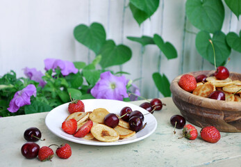 Homemade Mini pancakes and fresh strawberries, cherries on an old wooden table. Garden flowers in the background.