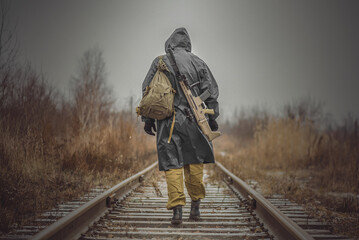 Soldier in raincoat with a backpack and rifle is walking away on the railroad. - obrazy, fototapety, plakaty