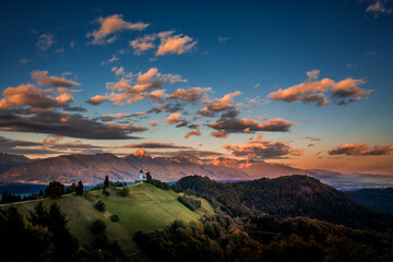 Famous landmark Jamnik church on hilltop. Green grassy slope in shadow. Alps mountains illuminated by evening sunset sun. Colorful sky at golden hour. Amazing Slovenia nature landscape