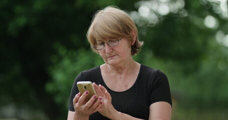 Senior concentrated woman in glasses writing message on her brand-new smartphone. standing on the street green tree background Straight shot