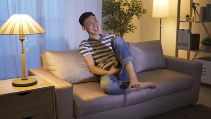 Obraz premium laid-back holiday asian guy changing tv channels found stand-up comedy and can't stop raising leg while laughing. korean male couch potato enjoying hilarious series at home alone before bedtime.
