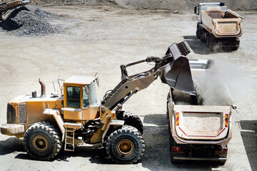 wheel loader with scoop working on construction site and loading gravel on dumper trucks. Heavy duty machinery working