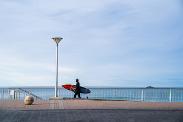 Surfer wearing wetsuit with surfboard watching ocean waves crash over rocks at St. Clair beach,...