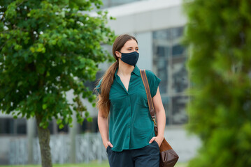 A woman in a medical face mask walks between trees thrusting hands into pockets of trousers in downtown. A girl keeping social distance wears a protective face mask to avoid the spread of coronavirus