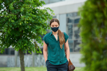 Sad woman in a medical face mask walks between trees thrusting hands into pockets of trousers in downtown. Girl keeping social distance wears a protective face mask to avoid the spread of coronavirus