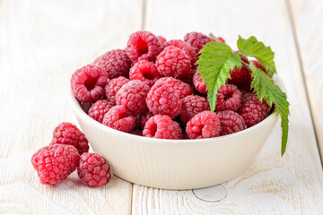 Whole ripe sweet raspberries and green leaf in a beige ceramic bowl on a white wood table. Tasty fresh organic fruits and berries, vegetarian and healthy eating. Raw food diet.