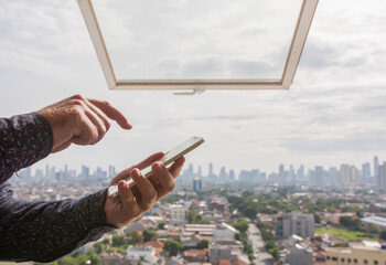 A man types a message on a smartphone against the background of an open window and a panorama of the city of Jakarta. Hands close up.