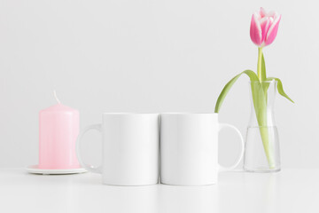 Two mugs mockup with pink tulips in a vase and candle on a white table.