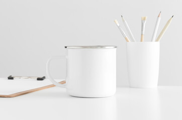 Enamel mug mockup with workspace accessories on a white table.