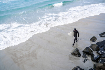 Surfer wearing wetsuit with surfboard watching ocean waves crash over rocks at St. Clair beach, Dunedin, New Zealand.