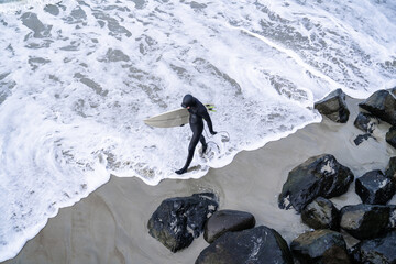 Surfer wearing wetsuit with surfboard watching ocean waves crash over rocks at St. Clair beach, Dunedin, New Zealand.
