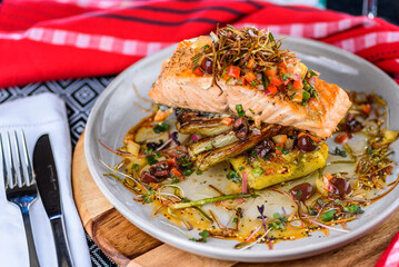 A seafood dinner serve of a piece of Salmon on top of grilled vegetables, in a round white plate