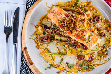 A seafood dinner serve of a piece of Salmon on top of grilled vegetables, in a round white plate
