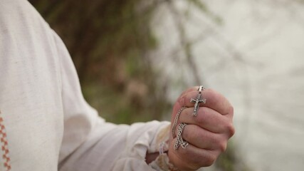 Old grandfather senior elderly man standing on the river bank in an embroidered shirt is conceived, remembers, recalling past and kisses silver cross with a chain. Close up