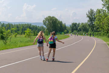 Fototapeta premium Two girls are rollerblading holding hands. Asphalt walkway in park, sunny, green trees. View from the back. Long hair, backpacks, short shorts. Concept of friendship, love, vacation. 