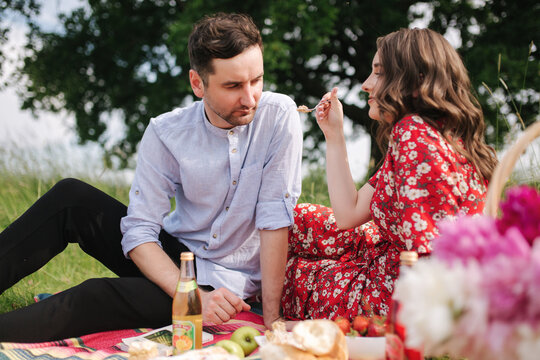 Happy couple spend time outdoors on mini picnic. Attractive woman and handsome man sits on red blanker by the big tree