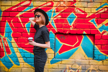 Pretty young smiling girl with cup of coffee in black hat and sunglasses stand against brick wall with colorful graffiti
