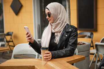 modern stylish muslim woman in hijab, leather jacket and black abaya sitting in city street cafe using mobile phone in sunglasses, drinking coffee