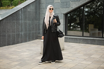 modern stylish muslim woman in hijab, leather jacket and black abaya walking in city street ,...