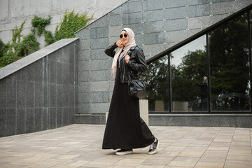 modern stylish muslim woman in hijab, leather jacket and black abaya walking in city street ,...