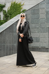 modern stylish muslim woman in hijab, leather jacket and black abaya walking in city street , wearing sunglasses and bag, fashion style trend - obrazy, fototapety, plakaty