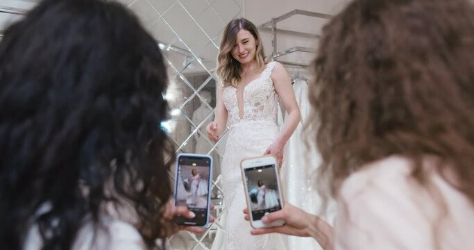 Happy bridesmaids taking pictures of beautiful blonde girl in wedding dress using smartphones. Bride posing for photo, dancing, smiling in dress on hanger in front of mirror in wedding salon.