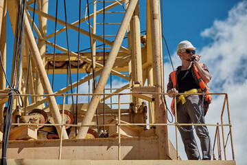 A construction engineer in a checked shirt, orange vest, and white hard hat on a yellow construction crane with a walkie-talkie in his hands. foreman, supervisor, worker