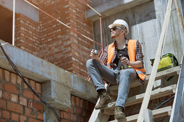 A civil engineer in a checked shirt, orange vest, and white hard hat. Happy young engineer, architect, worker sitting on a construction site and resting at lunch