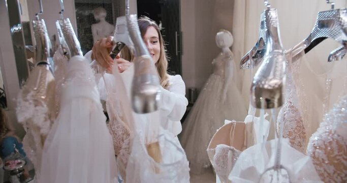 Close up portait of blonde bride searching for dress in wedding salon. Wedding dresses hang on hangers. A lot of dresses. Preparing marriage, shopping with girlfriends concept.