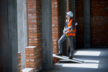 A construction engineer in a checked shirt orange vest and white on a construction site with a drawing in his hands