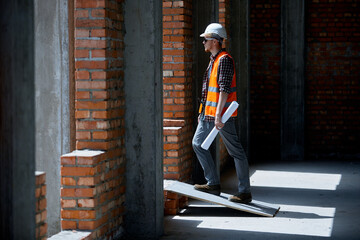 A construction engineer in a checked shirt orange vest and white on a construction site with a drawing in his hands