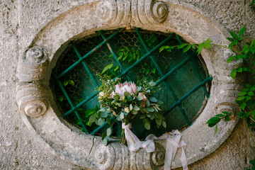 bridal bouquet of pink protea, branches of eucalyptus tree, pink roses, capsella and white ribbons on the oval window of an ancient house