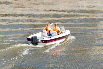 Young couple driving the speed boat in the river, wearing life vest. Shot at bright summer day. Nice tourist attraction to explore nature and the city.