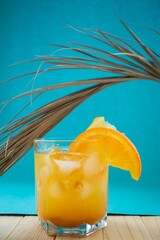 Summer orange-lemon cocktail with ice and alcohol on a blue and wooden background with a palm leaf. Tropical