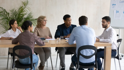 Interested diverse employees team listening to team leader mentor at meeting, sitting at table in...